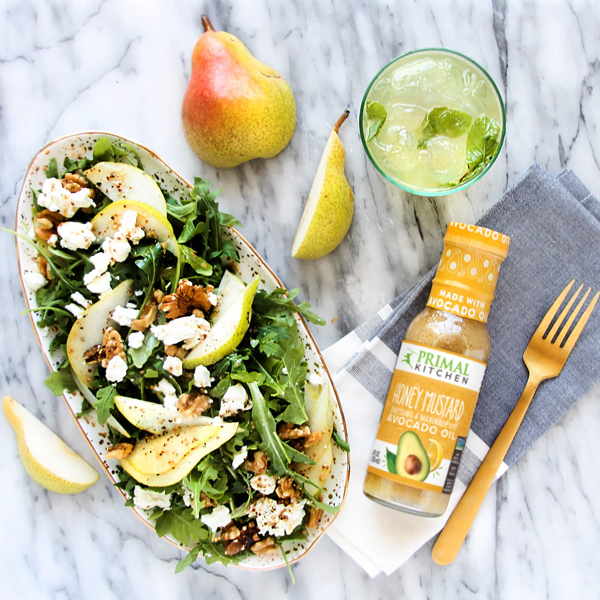 A pear and walnut salad with crumbled feta  in a bowl, next  to a bottle of Primal Kitchen Honey Mustard Dressing. Napkin, fork, drink, and sliced pear in the background.