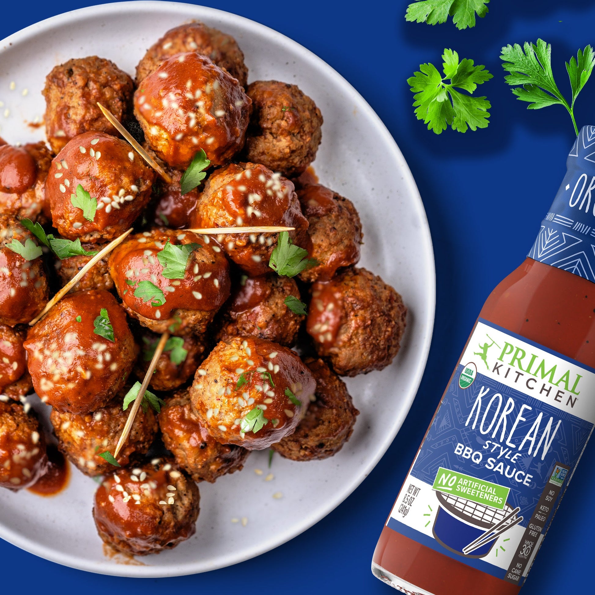 White plate with stacked beef meatballs topped with Korean BBQ Sauce, sesame seeds, and parsley with toothpicks, on a dark blue background with a bottle of Primal Kitchen Korean Style BBQ Sauce